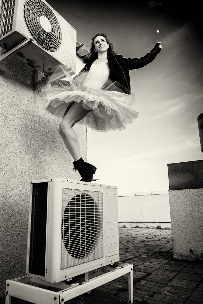 a ballerina in tutu, jacket and boots climbed on the air conditioner and poses against the sky von Alexandr